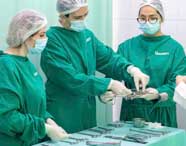 Grupo de estudantes de Enfermagem da Unoeste, usando aventais verdes e toucas, manuseia instrumentos cirúrgicos em sala de simulação. Eles estão reunidos ao redor de uma mesa metálica, praticando técnicas com pinças e outros equipamentos sob orientação.