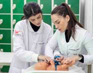 Duas alunas de Enfermagem, com jalecos brancos, realizam simulação de atendimento neonatal utilizando manequim de bebê. Uma mede o perímetro cefálico com fita azul enquanto a outra auxilia, em laboratório equipado com armários verdes ao fundo.