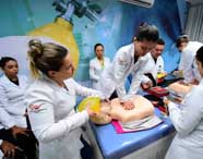 Estudantes de Enfermagem treinam reanimação cardiopulmonar em manequim adulto. Uma pessoa faz compressões torácicas, outra ventila com ambu amarelo e colegas acompanham o procedimento, todos em jaleco branco, simulando situação de parada cardiorrespiratória.