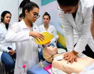 Alunos de Enfermagem praticam ventilação artificial em manequim de tórax. Um estudante insere tubo orotraqueal enquanto outro utiliza ambu. Todos usam jaleco branco, touca e luvas, em laboratório equipado com cartazes e materiais de simulação clínica.