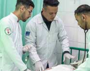 Estudantes de Enfermagem da Unoeste, em jaleco branco, realizam intubação orotraqueal em manequim adulto. Um aluno segura o tubo enquanto outro orienta, ambos com crachá e touca. Ao fundo, mais colegas participam da atividade prática no laboratório.