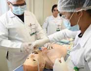 Estudantes do curso de Enfermagem realizam procedimento de intubação orotraqueal em manequim adulto sobre bancada. Usam jalecos brancos, luvas e toucas, com materiais médicos organizados ao redor. No crachá, é possível identificar a instituição Unoeste.