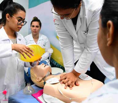 Estudantes do curso de Enfermagem realizam treinamento prático de reanimação cardiopulmonar em manequim adulto. Uma pessoa faz compressões torácicas enquanto outra utiliza um ambu amarelo para ventilação. Todos vestem jalecos brancos, simulando atendimento de emergência em ambiente acadêmico.