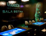 Sala Betha da Unoeste, ambiente moderno com mesas digitais interativas iluminadas em azul e cadeiras pretas. Parede preta exibe logotipo da universidade e mensagem de boas-vindas, espaço inovador para estudos em Engenharia Ambiental e Sanitária.