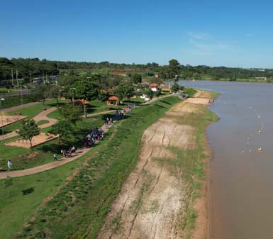 Vista aérea de margem de rio com vegetação, faixa de areia e gramado. À esquerda, parque arborizado com trilhas sinuosas, quadras de areia, quiosques e grupos de pessoas caminhando. O cenário ilustra atividades de campo típicas do curso de Engenharia Ambiental e Sanitária.