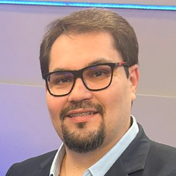 Aparecido, egresso do curso de jornalismo, em um ambiente interno com iluminação azul ao fundo. Ele veste um blazer preto e camisa clara, compondo um visual formal.