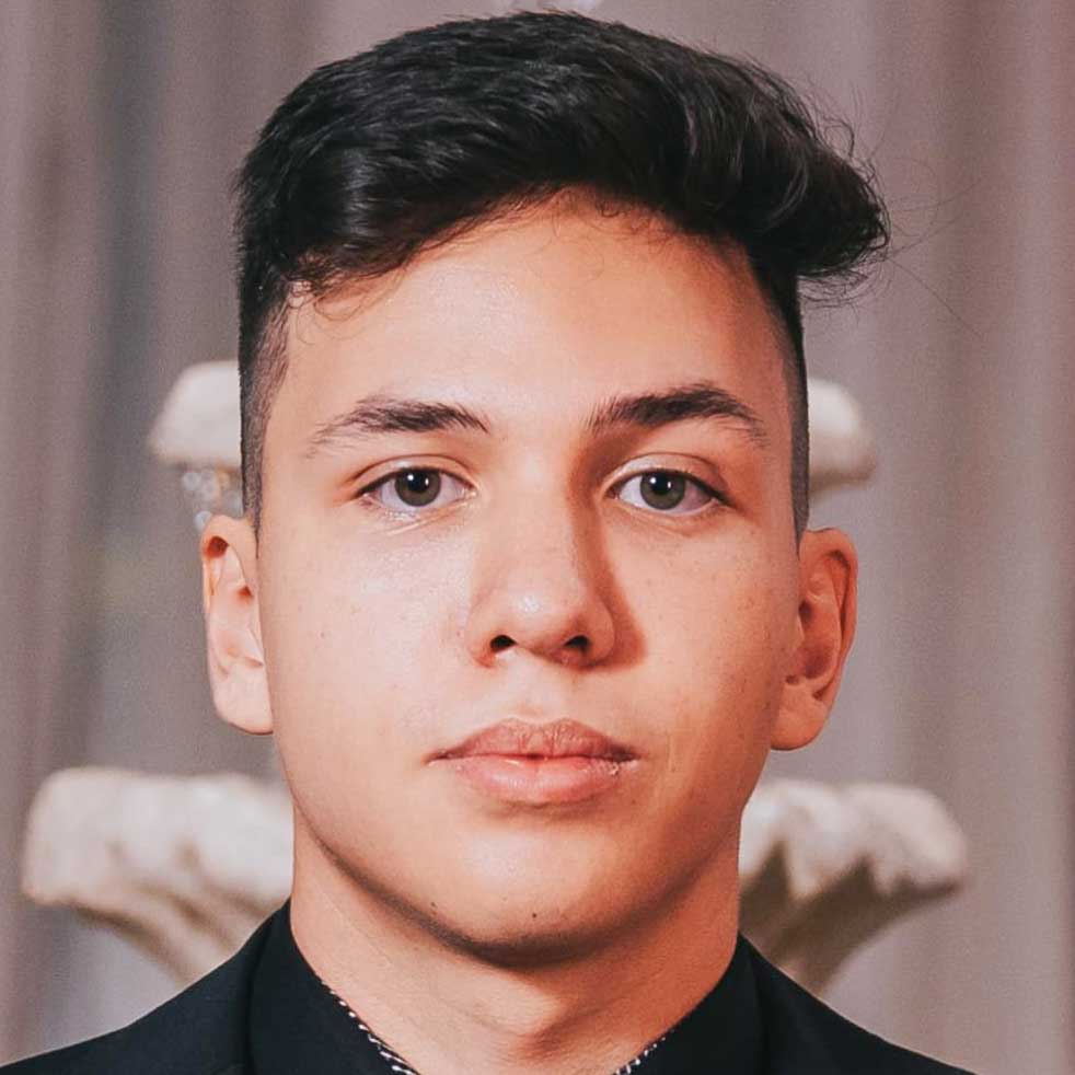 Enzzo, aluno do curso de jornalismo, posando em um ambiente interno com cortinas neutras e uma fonte decorativa ao fundo. Ele veste um terno preto, criando uma composição elegante.