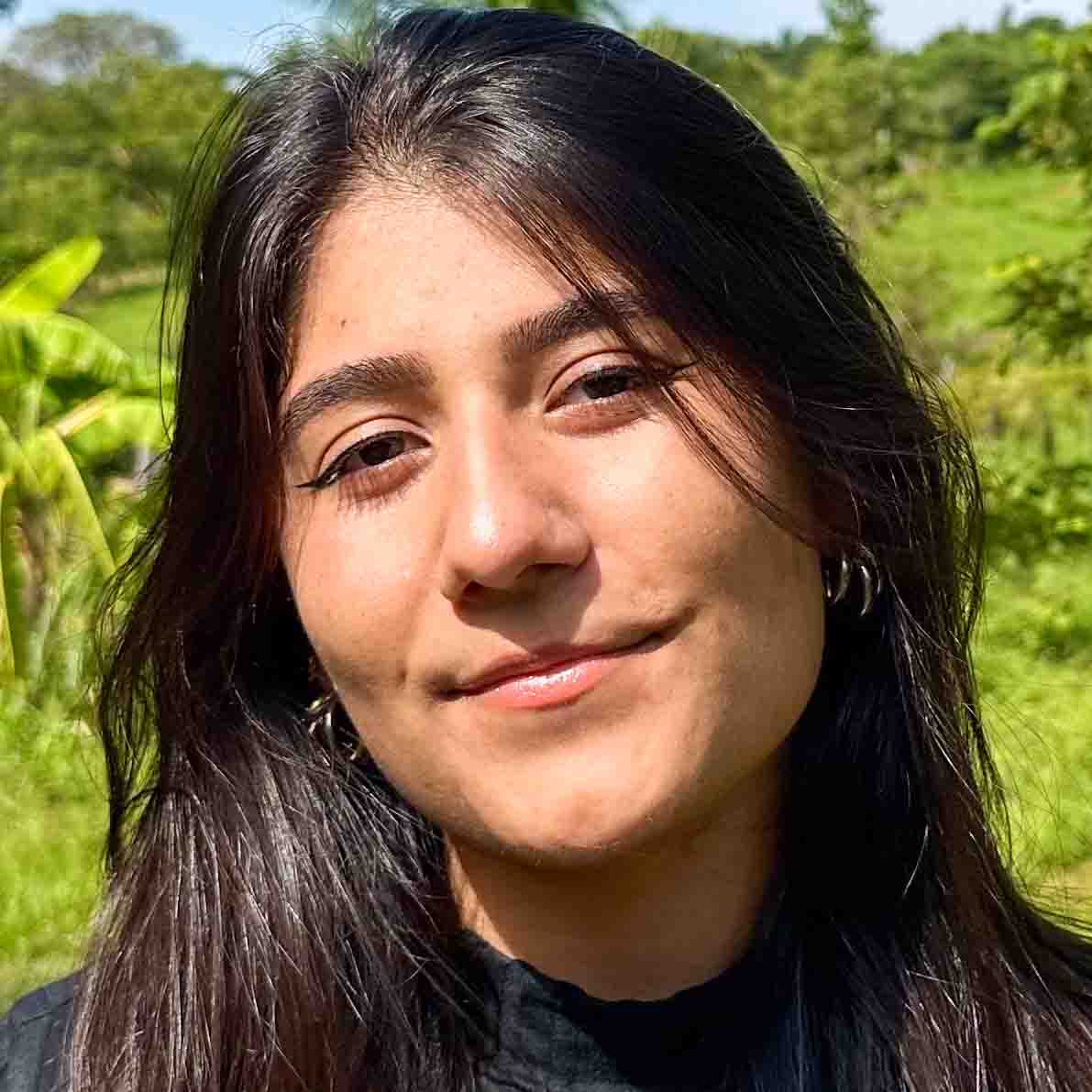Maria Clara, aluna do curso de jornalismo, em um ambiente externo com vegetação verde ao fundo. Ela veste uma blusa preta, complementando o cenário natural.
