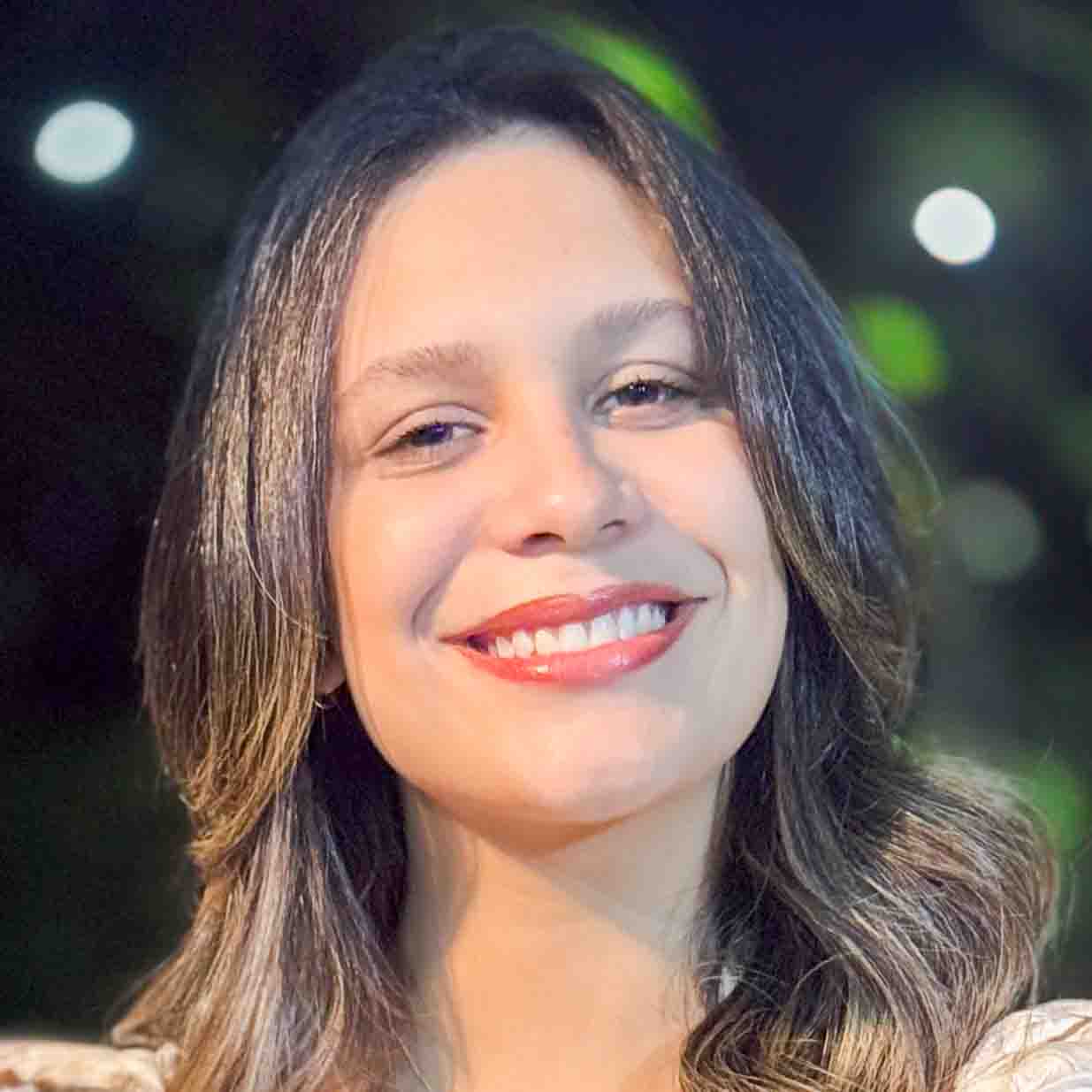 Marianne, aluna do curso de jornalismo, em um ambiente noturno com luzes desfocadas e vegetação ao fundo. Seu cabelo ondulado e bem cuidado destaca-se na composição iluminada.