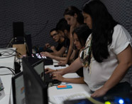Vários alunos de jornalismo trabalham lado a lado em laboratório de TV, utilizando computadores e mesas de controle. Uma mulher de cabelos longos e camisa branca orienta colegas, demonstrando integração entre teoria e prática no ensino superior, com paredes revestidas para isolamento acústico.