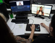 Dois estudantes de jornalismo editam vídeo em dupla, sentados frente a dois monitores. Um monitor exibe software de edição, o outro mostra entrevista de mulher com blusa clara. Na mesa, teclado, mousepad, anotações e caneta destacam o processo detalhado de produção audiovisual acadêmica.