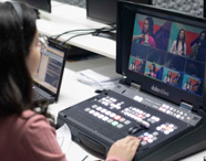 Mão de estudante ajusta mesa de som profissional com diversos botões e knobs coloridos, enquanto outro aluno ao fundo utiliza computador. O equipamento é fundamental para atividades práticas do curso de jornalismo, mostrando a importância da tecnologia no aprendizado em laboratório.
