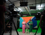 Estúdio de gravação com duas apresentadoras de jornalismo, uma de macacão rosa e outra verde, segurando roteiros. Elas estão em pé diante de fundo colorido geométrico, cercadas por câmeras, luzes e equipe técnica, ilustrando a vivência prática dos estudantes em telejornalismo.