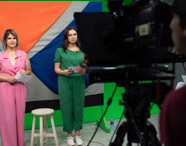 Duas estudantes apresentam telejornal em estúdio universitário, vestidas com macacão rosa e verde, segurando papéis. Ao redor, câmeras e iluminação profissional. Fundo geométrico colorido destaca o ambiente criativo e tecnológico do curso de jornalismo durante gravação prática.