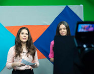 Duas estudantes apresentam programa de telejornal em estúdio, uma de blusa clara e outra de rosa, segurando roteiros. Câmera em primeiro plano aponta para elas, fundo decorado com formas geométricas coloridas, ambiente típico de laboratório de TV universitário.