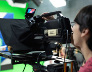 Estudante de jornalismo opera câmera profissional Sony com etiqueta “FACOPP CÂMERA 4”, focando apresentadoras ao fundo, uma de verde e outra de rosa. Ambiente de estúdio com fundo verde e painéis coloridos, ressaltando a infraestrutura e prática audiovisual do curso.