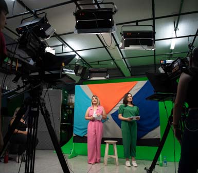 Estúdio de gravação com duas estudantes do curso de Jornalismo da Unoeste, uma vestindo macacão rosa e outra verde, em pé diante de fundo colorido sobre chroma key. Câmeras profissionais, iluminação de teto e tripés compõem o ambiente de produção audiovisual acadêmica.