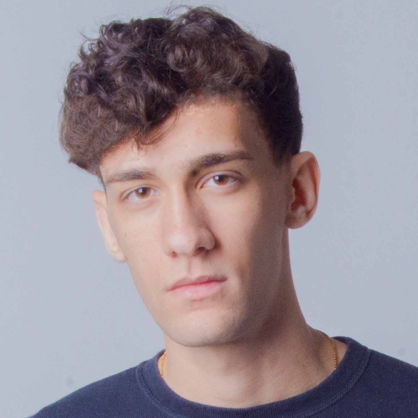Arthur, aluno de Publicidade e Propaganda, posando em um ambiente interno com um fundo claro. Ele tem cabelos cacheados e uma expressão séria.