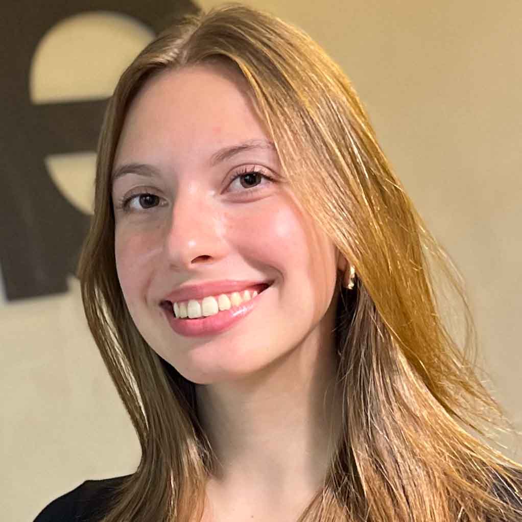 Giovanna, aluna de Publicidade e Propaganda, posando em um ambiente interno com um logotipo desfocado ao fundo. Ela tem cabelos longos e lisos, e usa brincos de argola, com um sorriso largo.