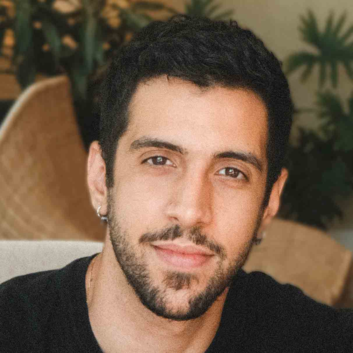 João, egresso de Publicidade e Propaganda, posando em um ambiente interno com plantas ao fundo. Ele tem barba e bigode, e usa brincos de argola, com uma expressão neutra.