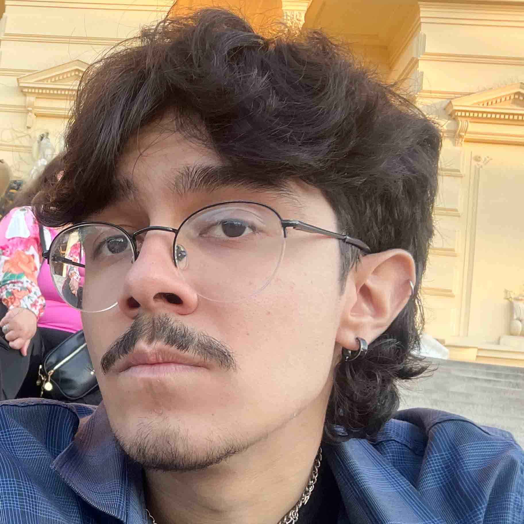 Leonardo, aluno de Publicidade e Propaganda, posando em um ambiente externo com um prédio histórico ao fundo. Ele usa óculos e tem bigode e barba rala, com cabelos compridos e uma expressão séria.