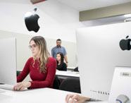 Alunos do curso de Publicidade e Propaganda participam de aula prática em laboratório equipado com computadores iMac. Em destaque, estudante de blusa vermelha opera o computador, enquanto outros alunos e o professor interagem ao fundo. Ambiente organizado, com mesas brancas e decoração minimalista, típico de universidades.