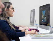 Estudantes de Publicidade e Propaganda utilizam computadores iMac em laboratório, focados em suas tarefas. A imagem mostra perfil lateral dos alunos, alguns com tatuagens e acessórios, destacando diversidade. O ambiente é claro, com mesas brancas e clima colaborativo, característico de instituições de ensino criativo.