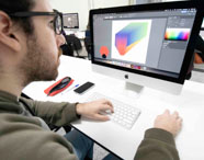Estudante do curso de Publicidade e Propaganda cria arte digital no Adobe Illustrator em um iMac. A tela exibe uma composição gráfica colorida. Sobre a mesa, caderno de capa florida e estojo listrado rosa e preto. Ambiente moderno, com iluminação clara e foco na criatividade e tecnologia.