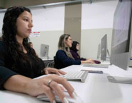 Turma de Publicidade e Propaganda realiza atividades em laboratório de informática, com computadores iMac alinhados sobre mesas brancas. Alunos digitam e usam mouse, demonstrando envolvimento em projetos digitais. O ambiente é bem iluminado, com paredes neutras e quadros informativos ao fundo.
