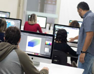 Estudante de Publicidade e Propaganda desenvolve projeto gráfico em iMac, com tela exibindo interface de design digital. Teclado branco, mouse e celular sobre a mesa. Ao fundo, colegas também trabalham em criações digitais, ilustrando o cotidiano prático do curso em ambiente universitário moderno.