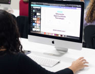 Professor orienta estudante de Publicidade e Propaganda durante produção de arte gráfica em laboratório de Macs. Ambos observam atentamente a tela do iMac, que exibe projeto visual. O ambiente é claro, com mesas brancas e outros alunos ao redor, reforçando o caráter interativo do ensino superior.