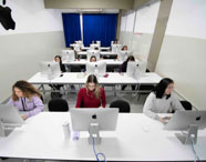 Vista aérea do laboratório de Macs da faculdade de Publicidade e Propaganda, mostrando três fileiras de mesas brancas com estudantes em frente aos iMacs. O ambiente é amplo, organizado e bem iluminado, com cortinas azuis ao fundo e atmosfera moderna, ideal para atividades práticas do curso.