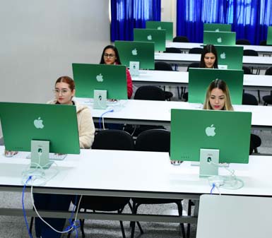 Laboratório de informática do curso de Publicidade e Propaganda da Unoeste Presidente Prudente, com estudantes sentados em frente a computadores iMac verdes organizados em fileiras. O ambiente é moderno, com mesas brancas, cadeiras pretas e cortinas azuis ao fundo, proporcionando espaço adequado para atividades criativas e digitais.
