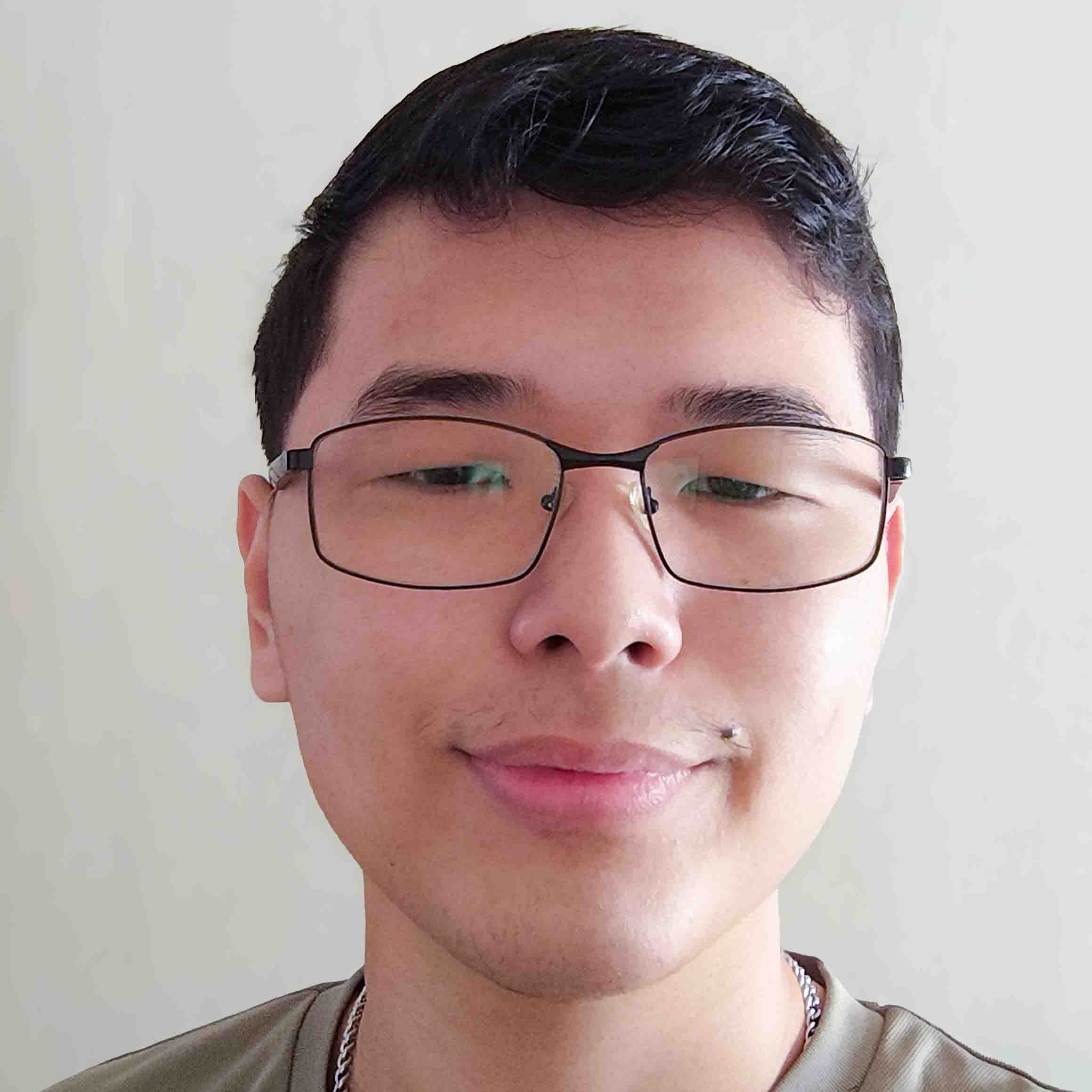 Adriano, aluno de Design Gráfico, posando em um ambiente interno com um fundo claro. Ele usa óculos de armação escura e tem um pequeno piercing no lábio superior, com uma expressão neutra.