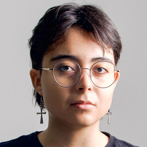 Letícia, aluna de Design Gráfico, posando em um ambiente interno com um fundo claro. Ela usa óculos redondos e brincos de cruz, com cabelos curtos e uma expressão séria.