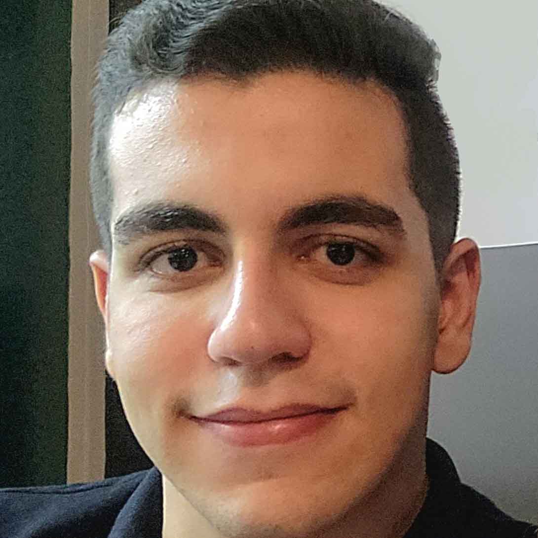 Matheus, aluno de Design Gráfico, posando em um ambiente interno com um fundo claro. Ele tem cabelos escuros e uma expressão amigável.