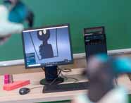Monitor exibe ilustração digital de personagem preto com olhos brancos, criada em software 3D. À frente, mão segura controle de realidade virtual. Na mesa, objetos geométricos coloridos e torre de computador Lenovo, em sala de aula do curso de Design Gráfico.