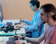 Dois estudantes sentados lado a lado em laboratório de informática, cada um usando mesa digitalizadora e caneta stylus diante de monitores. Sobre a mesa, óculos de realidade virtual Warrior. Atividade prática do curso de Design Gráfico, integrando desenho digital e tecnologia.