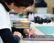 Estudante do curso de Design Gráfico desenha em tablet com caneta stylus, criando arte digital detalhada. Ela usa camiseta clara e acessórios, sentada à mesa com materiais artísticos e corte ao fundo. Ambiente universitário focado em práticas criativas digitais.