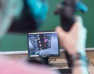 Pessoa interage com controles de realidade virtual HTC Vive diante de monitor Samsung que exibe modelagem 3D em ambiente gráfico. Sala equipada com computadores e materiais didáticos, típica do curso de Design Gráfico, onde inovação tecnológica é destaque.