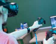 Pessoa interage com controles de realidade virtual HTC Vive diante de monitor Samsung que exibe modelagem 3D em ambiente gráfico. Sala equipada com computadores e materiais didáticos, típica do curso de Design Gráfico, onde inovação tecnológica é destaque.