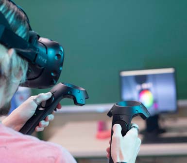 Estudante do curso de Design Gráfico em Prudente utiliza óculos de realidade virtual e dois controles nas mãos, interagindo com ambiente digital. Ao fundo, monitor exibe interface gráfica colorida. Sala de aula equipada, destacando tecnologia aplicada ao ensino de design na instituição.