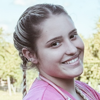 Anny, aluna do curso de veterinária, trajando um jaleco rosa e com um estetoscópio ao redor do pescoço. Ela está em um ambiente externo com fundo composto por árvores e céu azul, sugerindo um cenário rural ou de campo. A iluminação natural destaca os detalhes da vestimenta e reforça o contexto profissional e acadêmico.