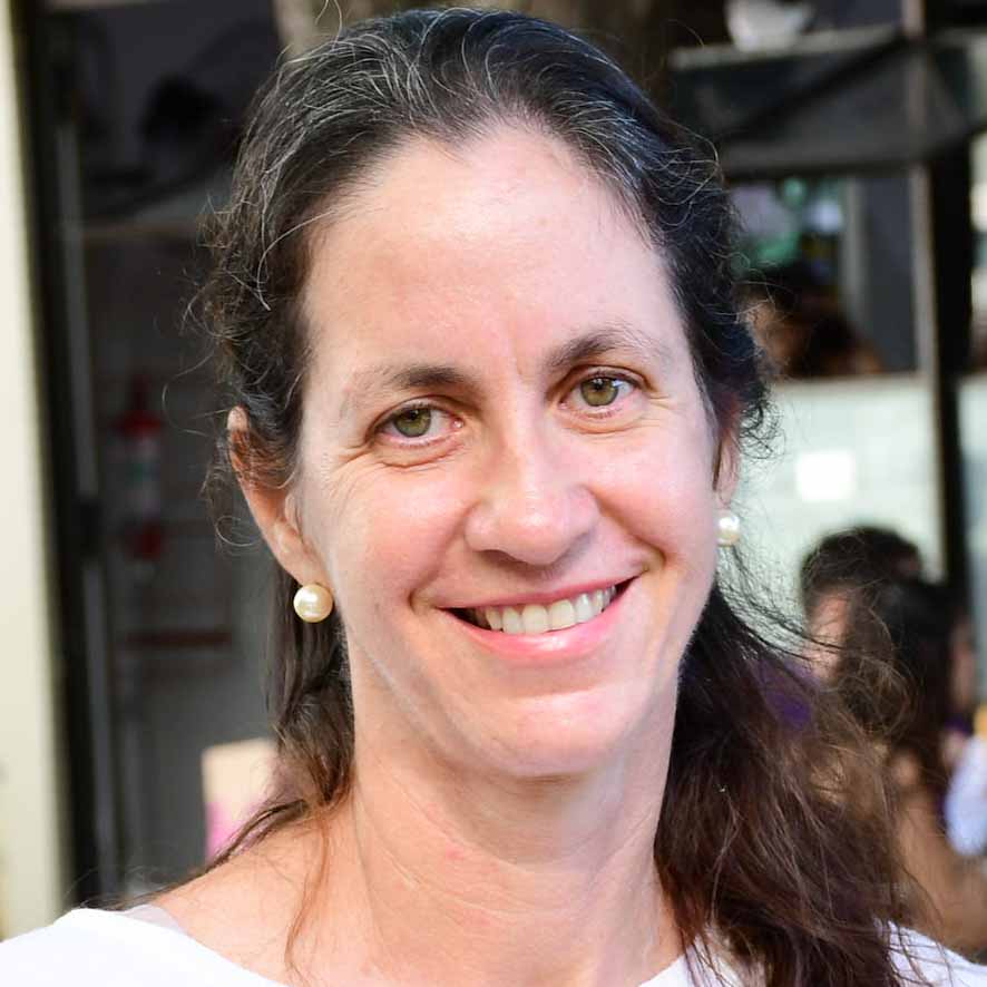 Cristiane, aluna do curso de veterinária, trajando uma camiseta branca. Ela está em um ambiente com fundo movimentado, incluindo pessoas e elementos arquitetônicos, sugerindo um evento ou atividade. A iluminação natural valoriza os detalhes da composição, criando um visual dinâmico e acolhedor.