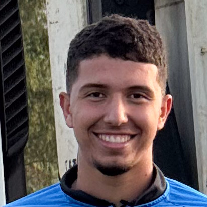 Carlos, aluno do curso de agronomia, trajando uma camisa azul. Ele está em um ambiente externo com fundo composto por estruturas metálicas e vegetação ao longe, sugerindo um cenário agrícola ou industrial. A iluminação natural destaca os detalhes da vestimenta e o contraste entre os elementos do fundo, reforçando o contexto prático e rural.