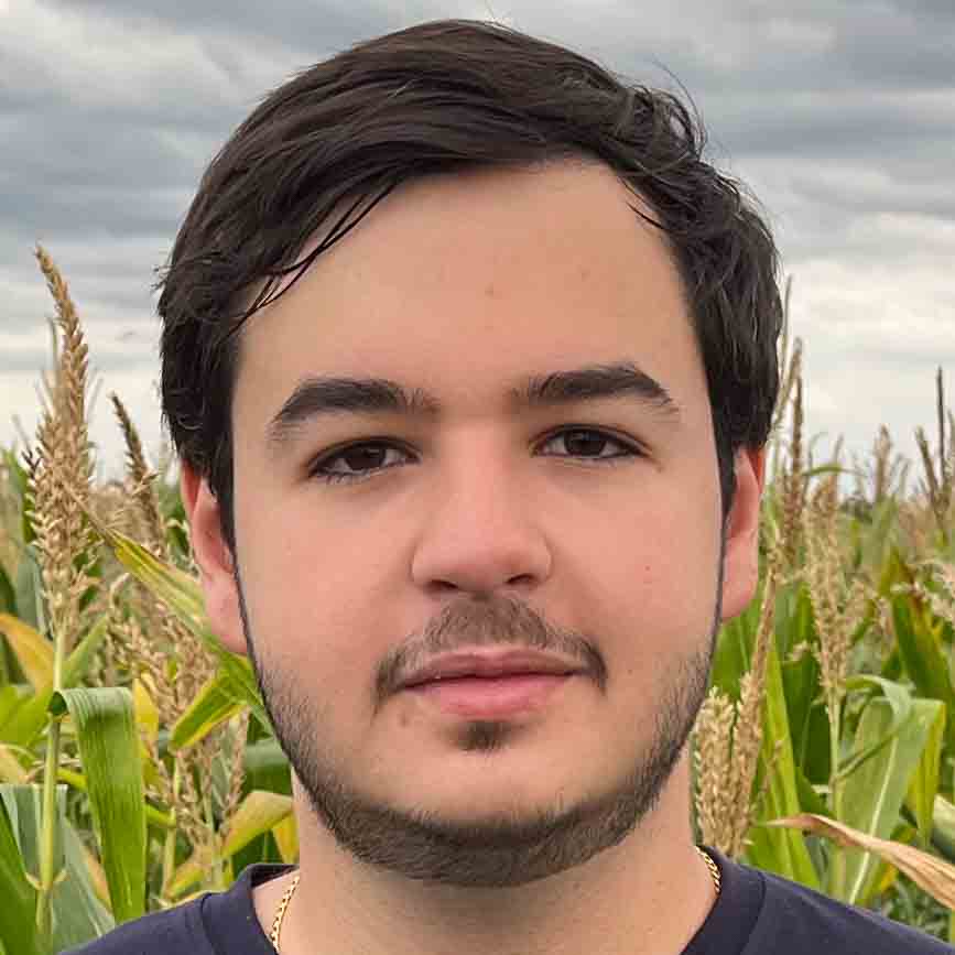 Gabriel, aluno do curso de agronomia, trajando uma camiseta preta. Ele está em um ambiente externo com fundo formado por uma plantação de milho sob um céu nublado, representando um cenário típico do campo. A iluminação difusa valoriza a composição natural, conectando o estudante ao ambiente agrícola.