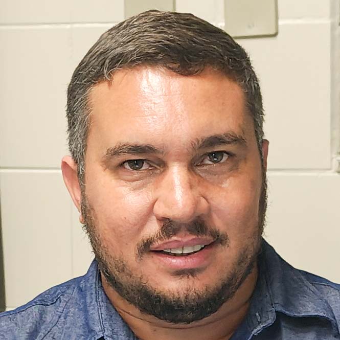 Luciano, egresso do curso de agronomia, trajando uma camisa jeans azul. Ele está em um ambiente interno claro, sugerindo um espaço técnico ou laboratório. A iluminação uniforme destaca os detalhes da roupa e cria um contraste simples e profissional, reforçando o contexto acadêmico e prático.