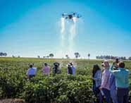 Alunos do curso de Agronomia em Presidente Prudente observam a demonstração de um drone agrícola em plantio, durante atividade prática em fazenda experimental. O drone pulveriza a plantação, enquanto estudantes acompanham a tecnologia no campo, reforçando inovação, tecnologia agrícola e aprendizado prático no agronegócio.