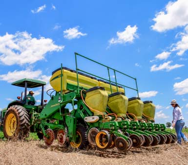 Perfil profissional agronomia Prudente: trator agrícola verde equipado com semeadora realiza plantio em campo aberto sob céu azul, acompanhado por dois profissionais de chapéu, que monitoram a operação. A imagem evidencia a aplicação de tecnologia, práticas sustentáveis e o aprendizado prático no campo, essenciais para a formação do engenheiro agrônomo no ambiente acadêmico e profissional.
