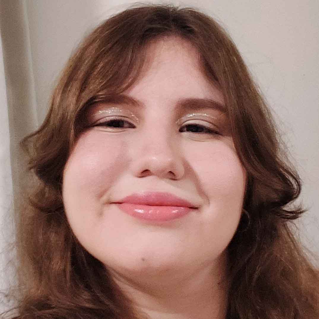 Clara, aluna de Design, posando em um ambiente interno com um fundo claro. Ela tem cabelos castanhos e usa maquiagem nos olhos, com um sorriso suave.